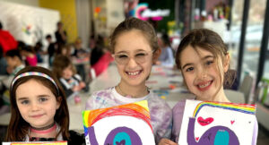 3 smiling children holding up colorful hand-made artwork