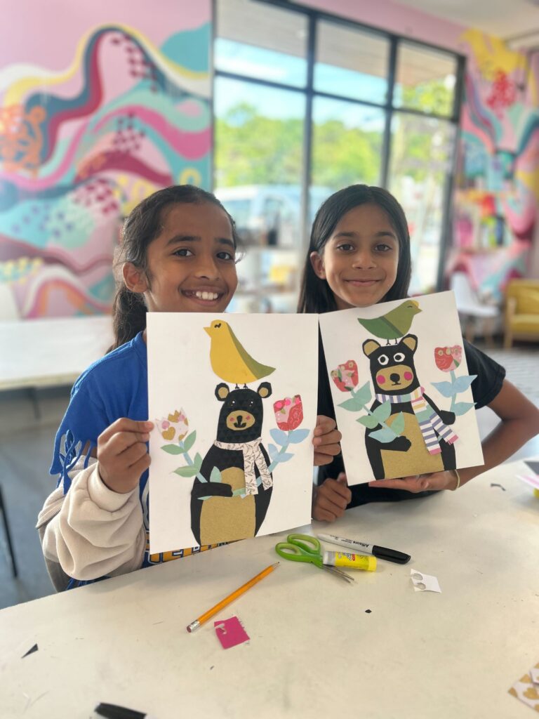 Two girls smiling, seated at a table and holding up their artwork.